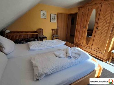 Das Schlafzimmer 1 der Ferienwohnung 5 ist im Landhausstil eingerichtet und bietet ein Doppelbett, ein Kinderbett, einen großen Kleiderschrank, Stühle zur Kleiderablage und einen Zugang zum Balkon. Idealer Ort für erholsame Tage im Harz.