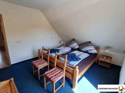 Das erste Schlafzimmer in Ferienwohnung 2 der Pension Quellenhof Altenau bietet ein Doppelbett, Kinderbett, 2 Nachttische, einen Kleiderschrank, 2 Stühle zur Kleiderablage und einen Balkonzugang.